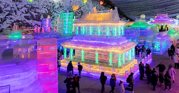 Nighttime ice sculpture park lit by neon rainbow lights; glowing ice buildings and towers with visitors walking among colorful, illuminated blocks.