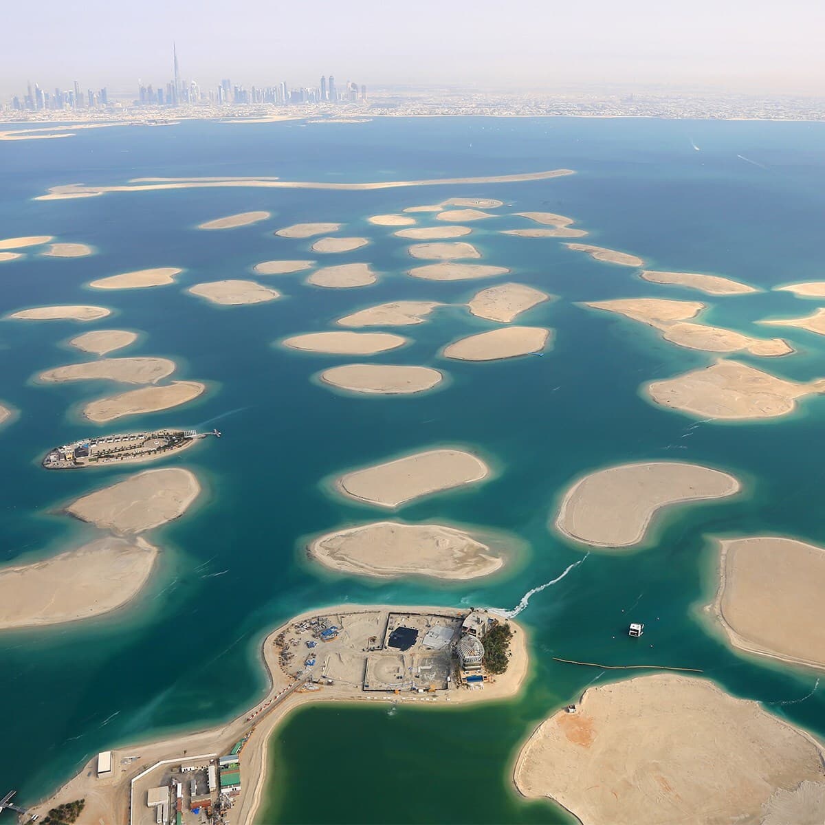 Aerial view of many sandy islands in turquoise water, with a distant city skyline; construction site on lower left and a small boat nearby.