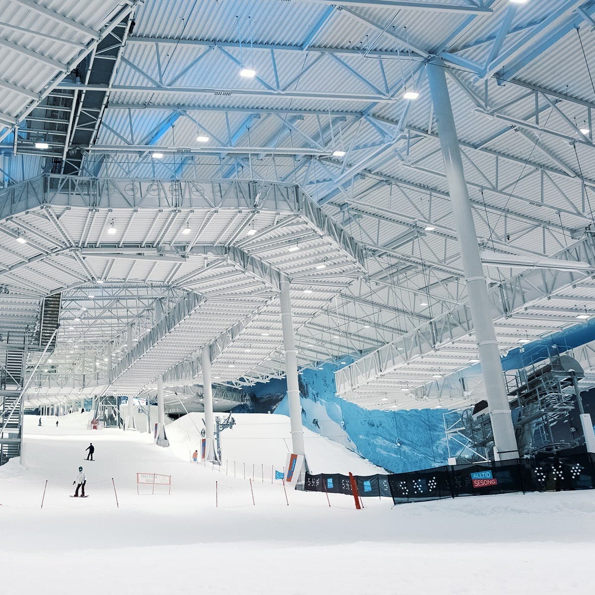 Indoor ski hall with a snow-covered slope, ramps and rails under a white steel-truss ceiling; several skiers glide along the slope near blue wall art and fencing.