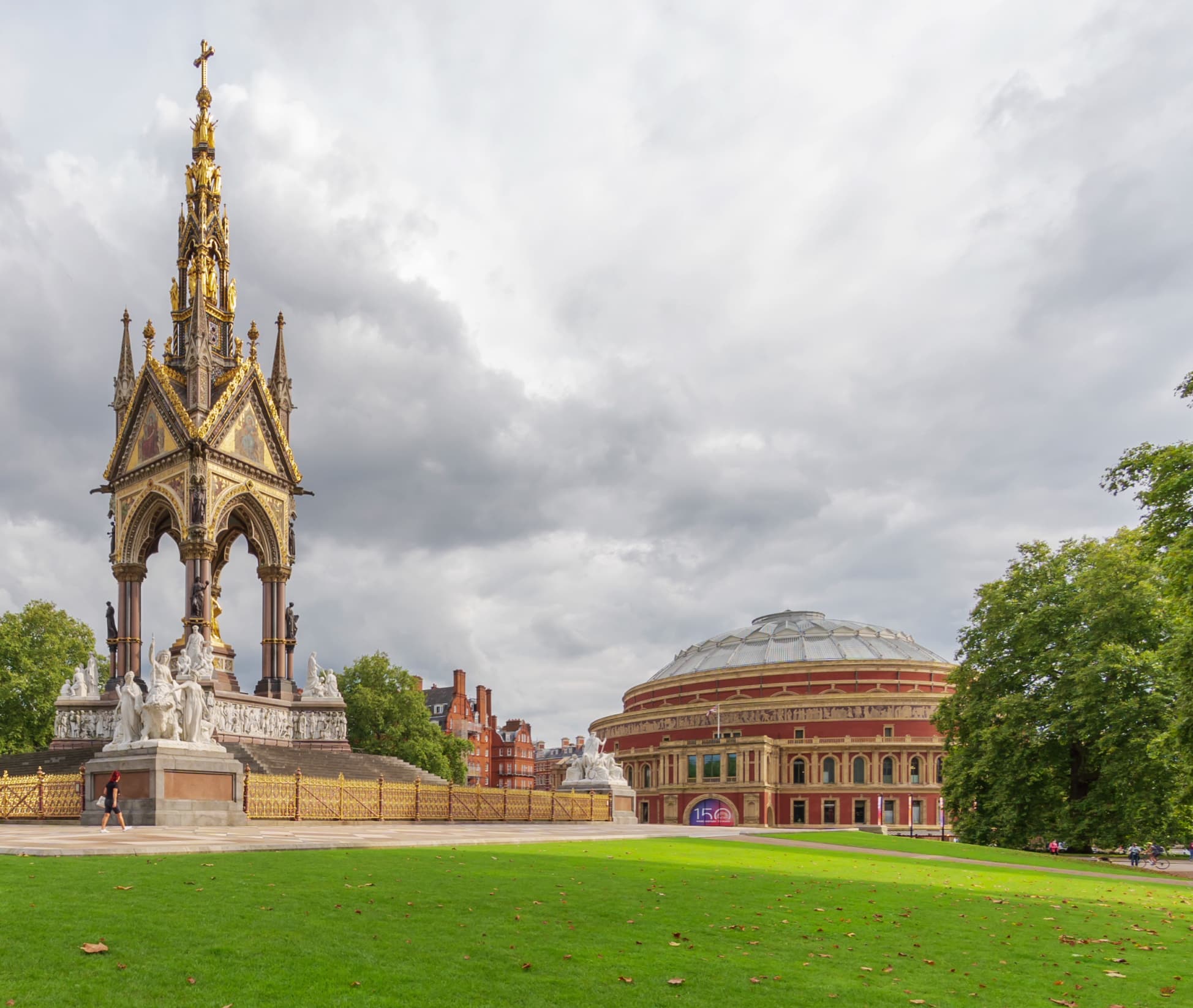 Ornate gilded spire monument with statues stands on a stone base in a green park, beside a circular red-brick domed building under a cloudy sky.