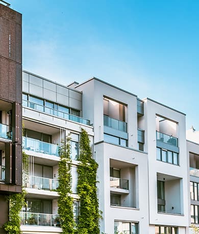 Modern white apartment building with glass balconies and an ivy-covered wall on the left, set against a bright blue sky.