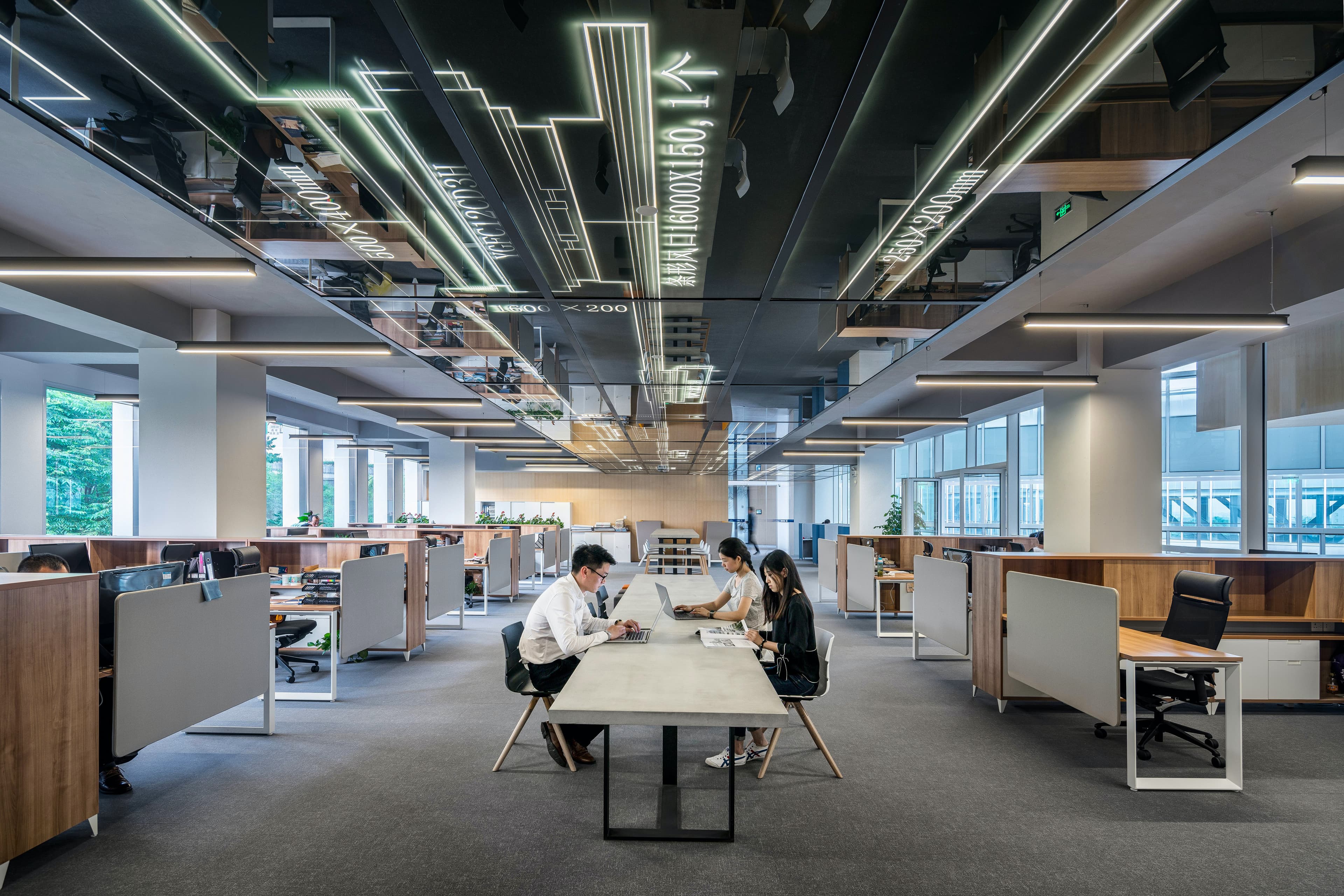 Three coworkers sit at a long central table in an open-plan office, surrounded by cubicles, large windows, and a neon-lined mirrored ceiling.