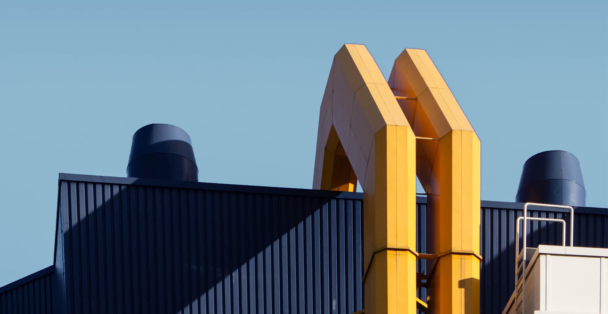 Blue metal building with dark vertical siding, a bold yellow double-arch sculpture on top, blue vents, and a white railing against a clear sky.