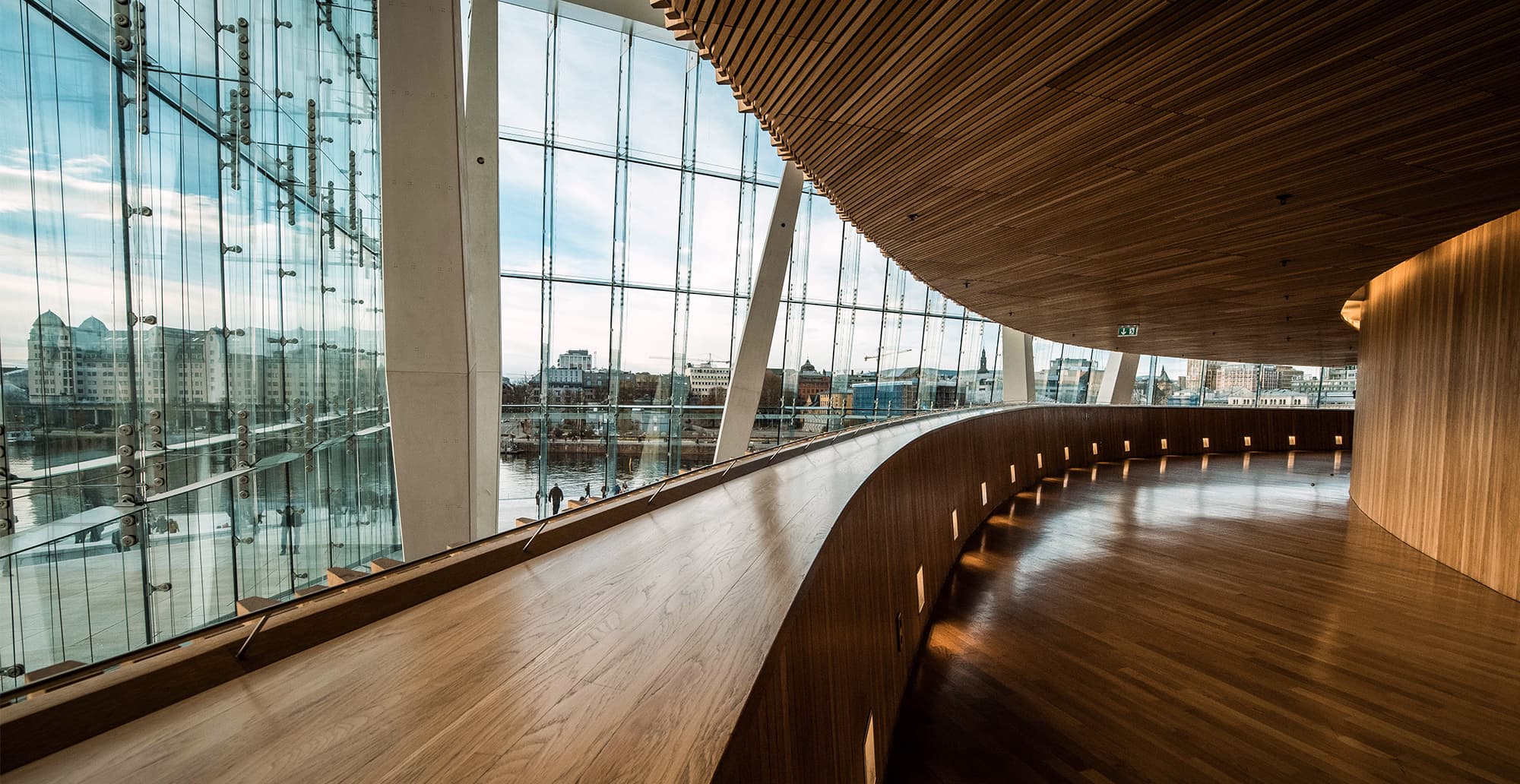 Curved wooden interior with a floor-to-ceiling glass wall; river and city skyline outside, warm lighting along the railing and ceiling.