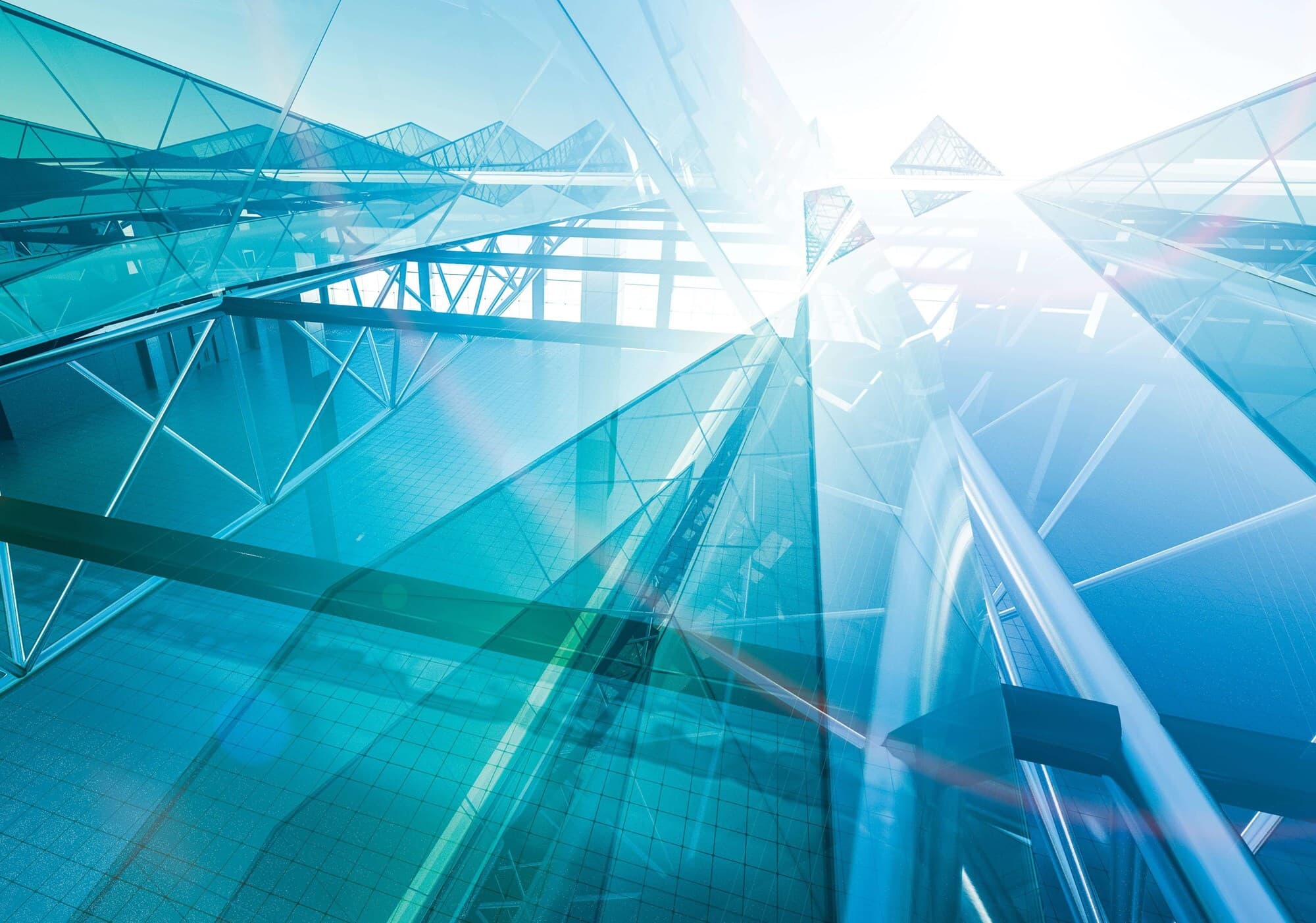 Blue glass atrium with crisscross metal framework, reflective walkways and bright light streaming through geometric glass panels.