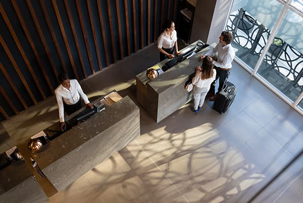 Top-down view of a hotel lobby with staff at two stone reception desks helping guests; luggage nearby, large windows, and patterned shadows on the tiled floor.
